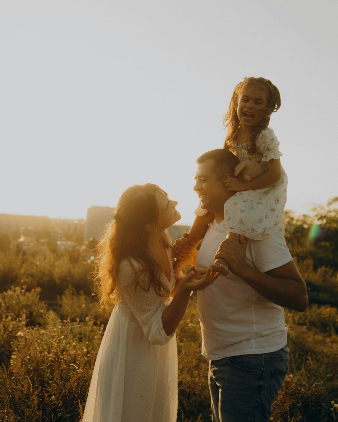 Pai com a filha nos ombros e a mãe estendendo as mãos para ela, ao pôr do sol